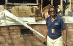 Woman standing in front of a fire-damaged brick building, wearing sunglasses, navy shirt, and a lanyard with an ID badge.