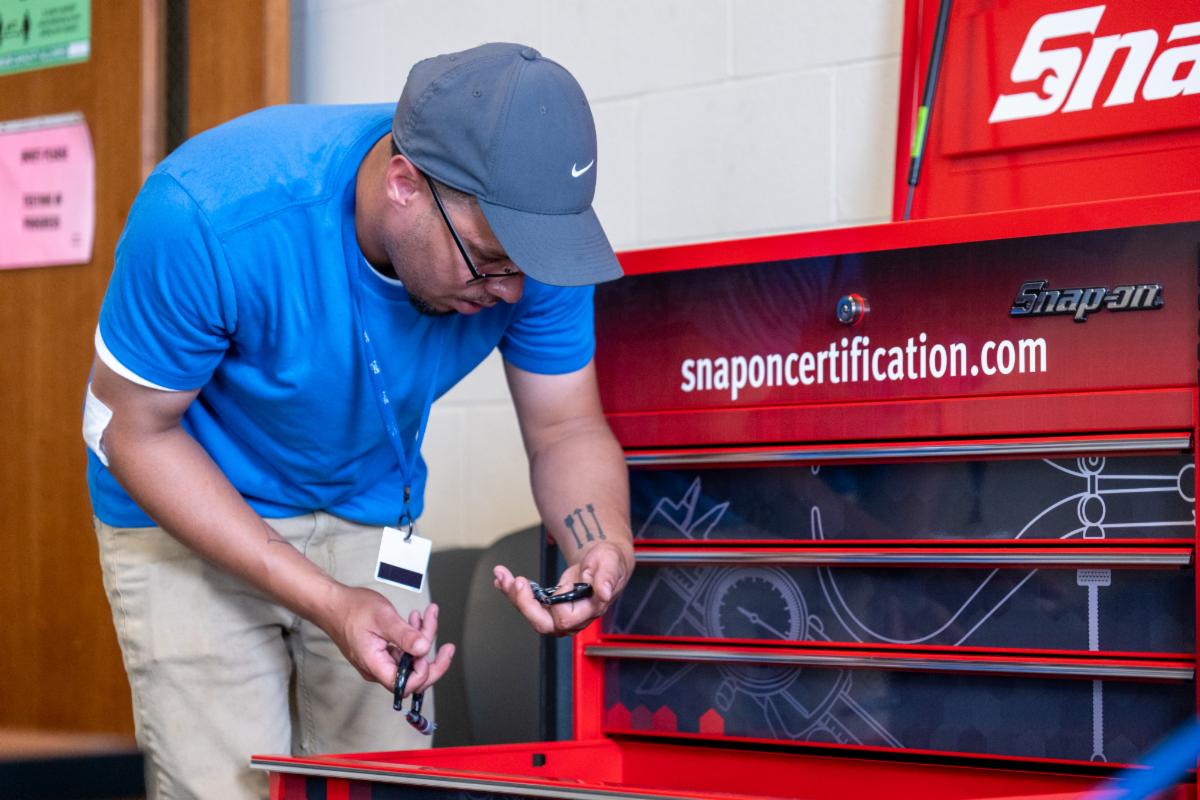 A man in a blue shirt works with tools near a red tool chest labeled "snaponcertification.com" in a workshop setting.