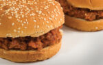A close-up of two Sloppy Joe sandwiches on sesame seed buns, filled with savory ground meat mixture, placed on a white surface.