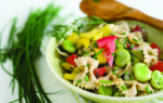 Bowl of pasta salad with farfalle, sliced yellow peppers, radishes, cucumbers, and fresh herbs, garnished with chives, next to a wooden spoon and chive bunch.