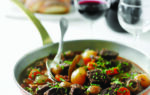 A pan of beef stew with vegetables, garnished with herbs, is served on a table with bread and two glasses of red wine in the background.