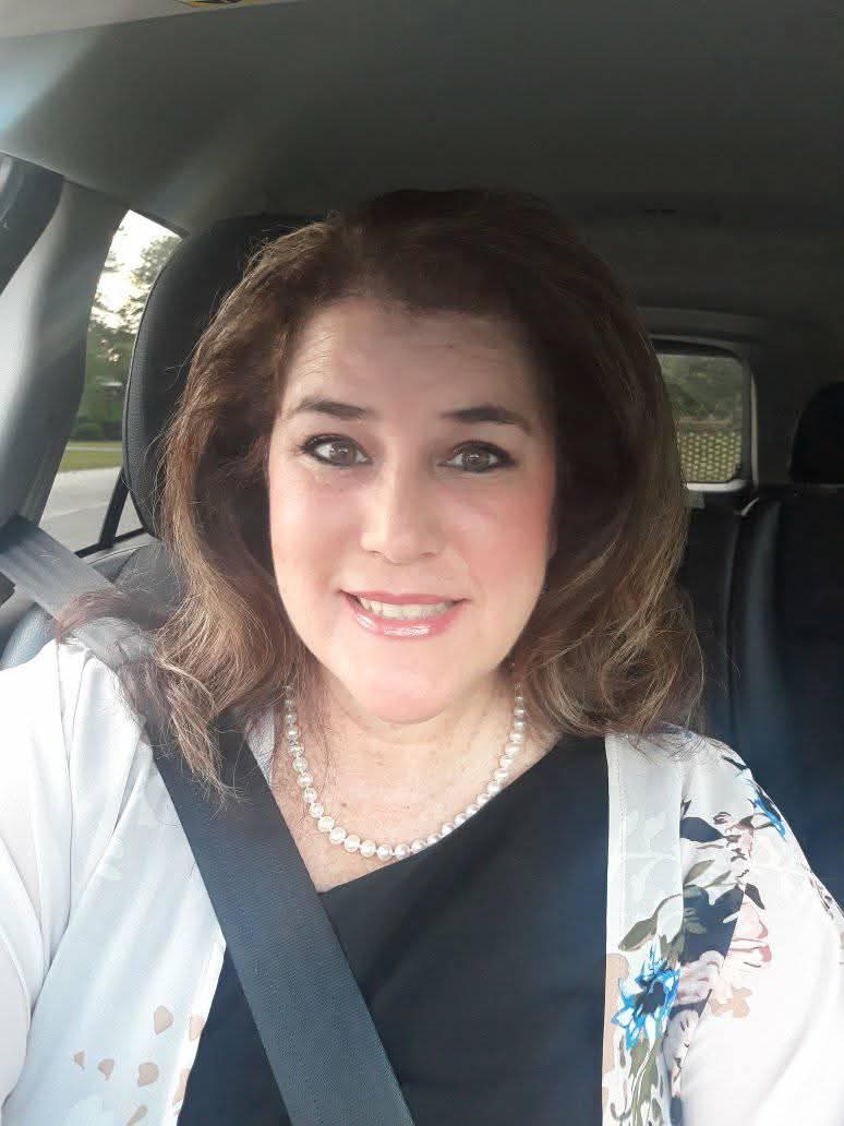Woman with shoulder-length brown hair smiles, sitting in a car, wearing a white top, black shirt, and a pearl necklace, seatbelt fastened.
