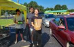 Three people standing in a parking lot near a red car, one holding a paper bag, with a yellow canopy and trees in the background.