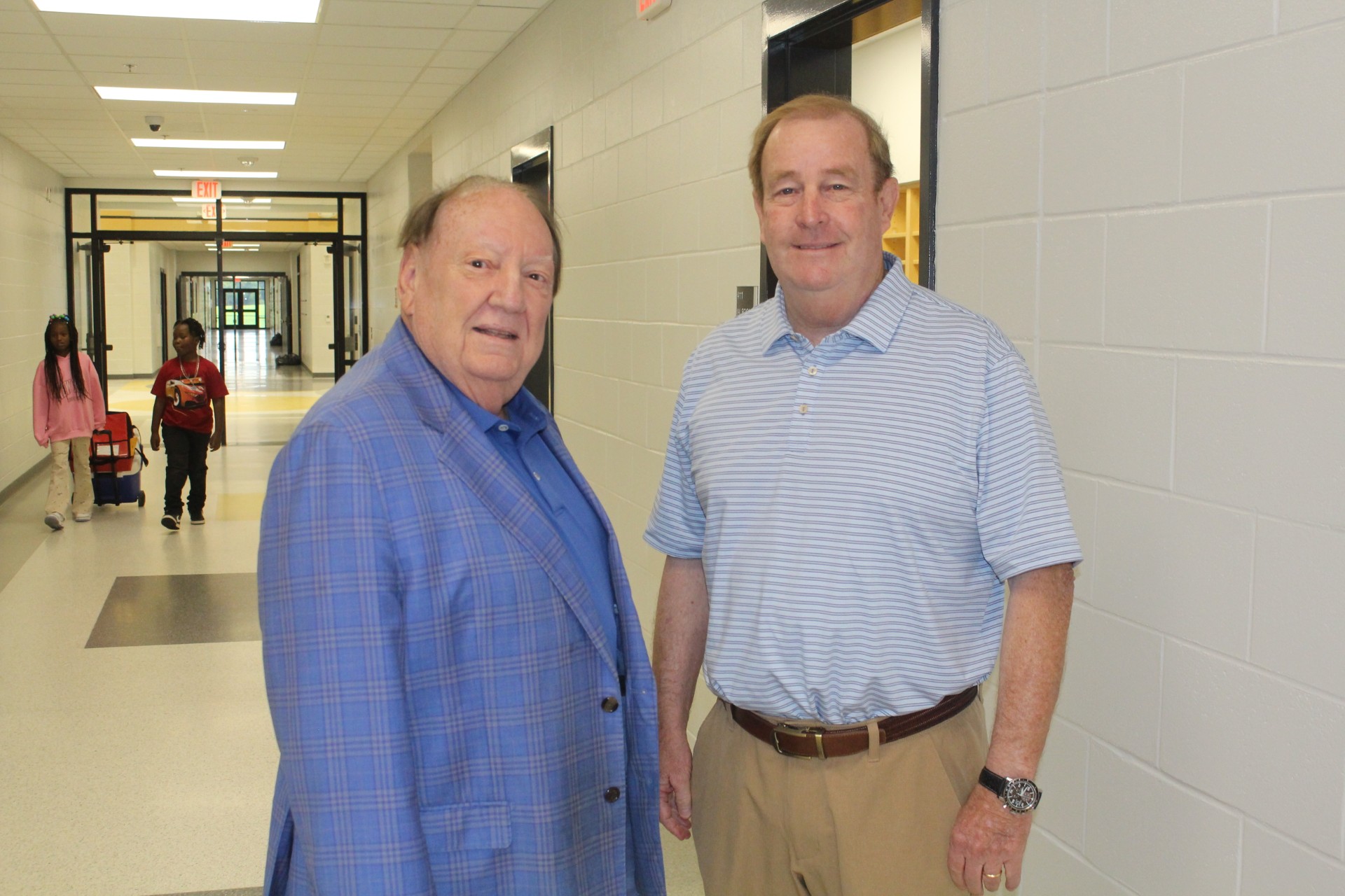 Two men standing in a hallway, one wearing a light blue blazer and the other in a light blue polo shirt, both smiling at the camera.