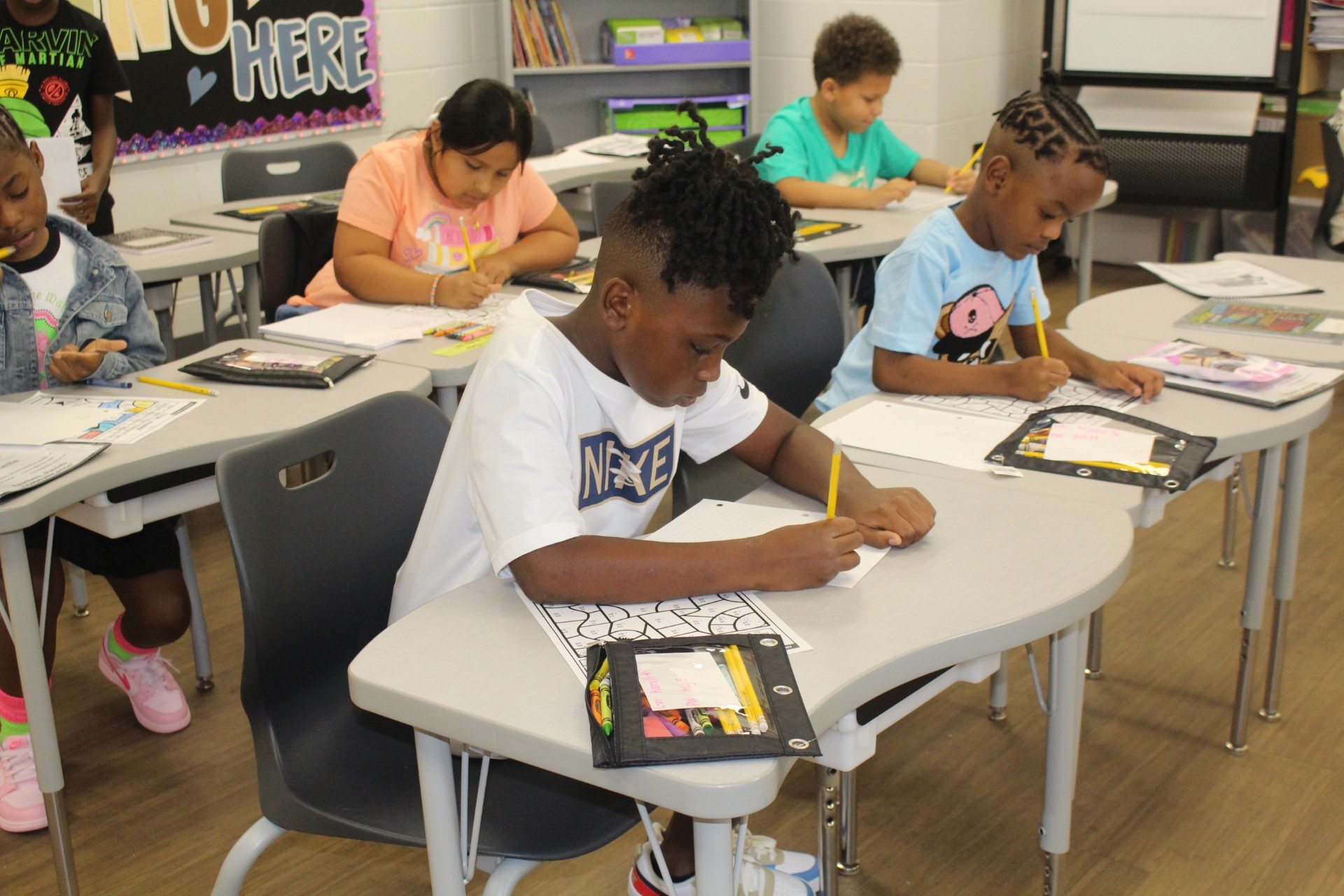 Four young students sit at desks in a classroom, focused and writing in notebooks during a lesson.