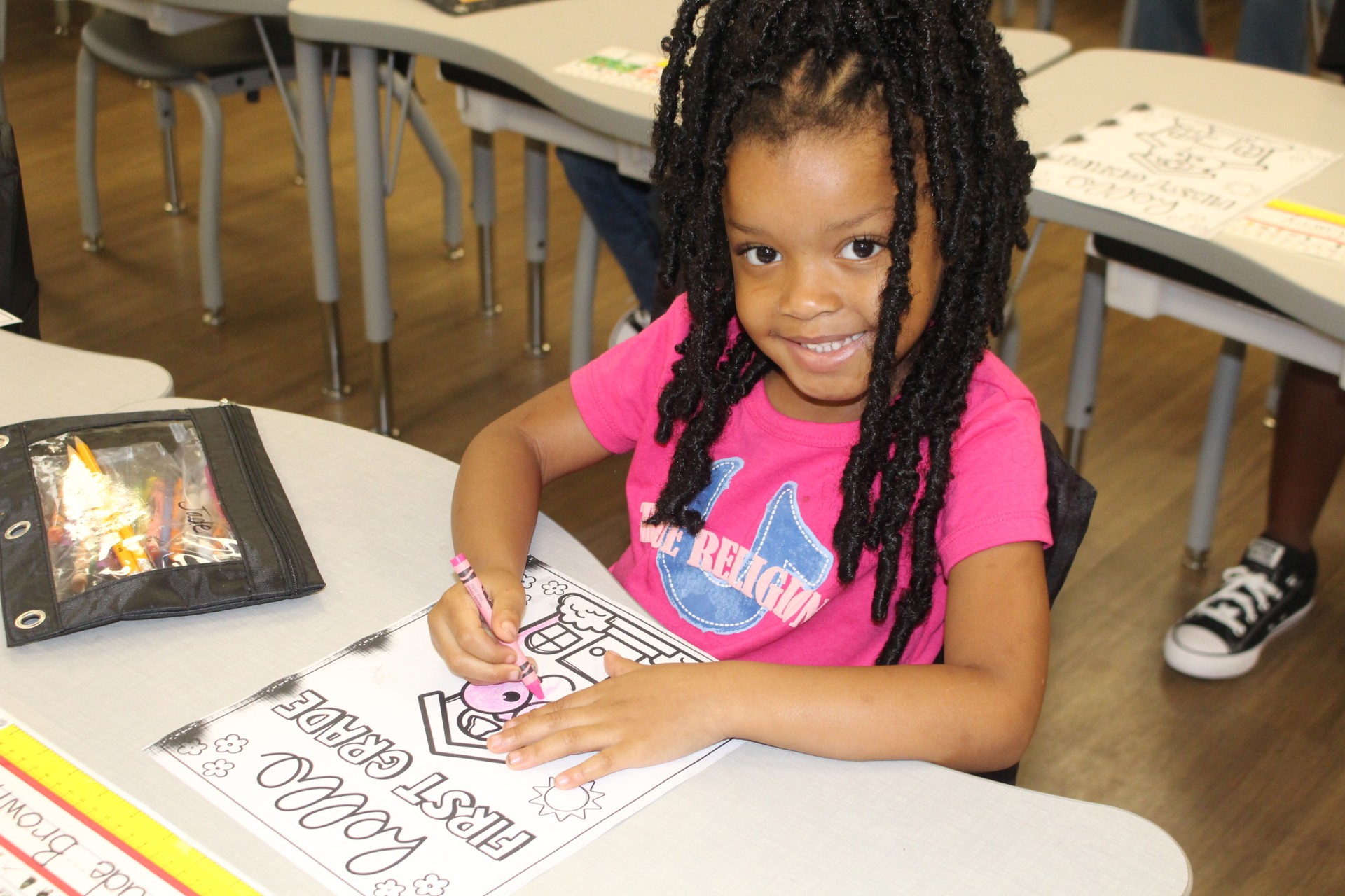 A young girl with braided hair colors a worksheet at her classroom desk, smiling at the camera while holding a crayon.