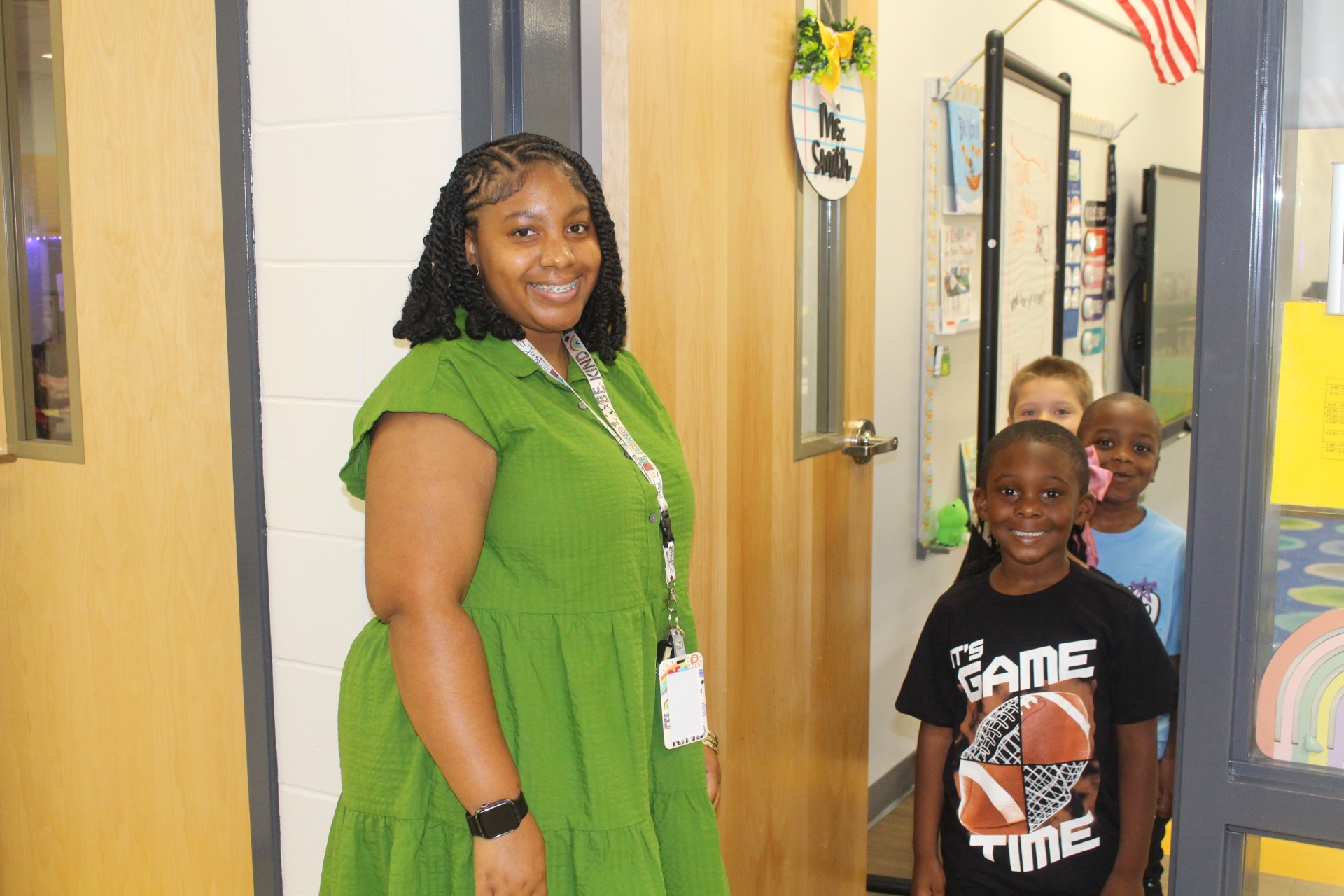 A woman in a green dress stands smiling by a classroom door, with several children smiling nearby in a school hallway.