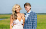 A smiling couple poses outdoors; the woman shows an engagement ring, wearing a white dress, while the man stands beside her in a blue plaid suit.