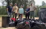 Five people stand outdoors behind a group of large filled trash bags, with trees in the background, participating in a cleanup activity.