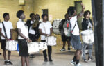 A group of young people in casual clothing practice playing drums indoors, some holding drumsticks, with backpacks and a brick wall in the background.