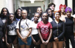 A group of twelve young people stand together in a hallway, smiling at the camera, dressed casually in t-shirts and shorts.