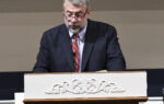 A man in a suit and red striped tie stands at a decorated podium, speaking or reading in a formal indoor setting.