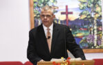 A man in a suit stands at a podium in a church, with a stained glass window and red cross in the background.