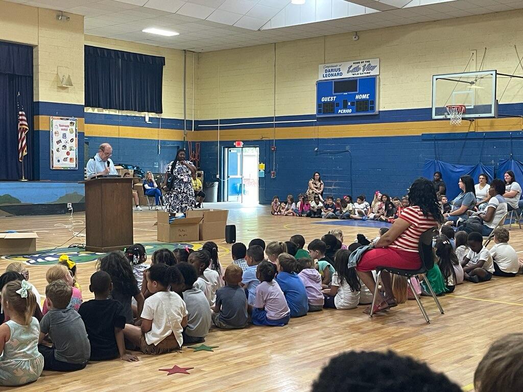 A group of children sit on a gym floor facing a speaker at a podium, with adults supervising in a school assembly setting.