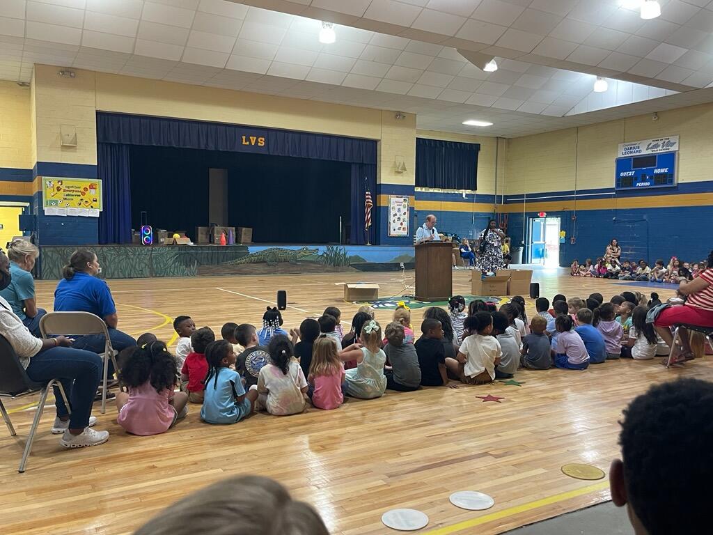 Children sit on a gym floor facing a stage where adults stand at a podium during a school assembly in a large indoor auditorium.