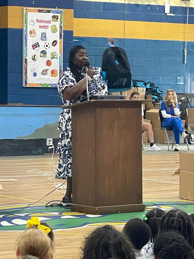 A woman speaks at a podium in a gymnasium, addressing an audience. People are seated in the background, and decorations are visible on the wall.