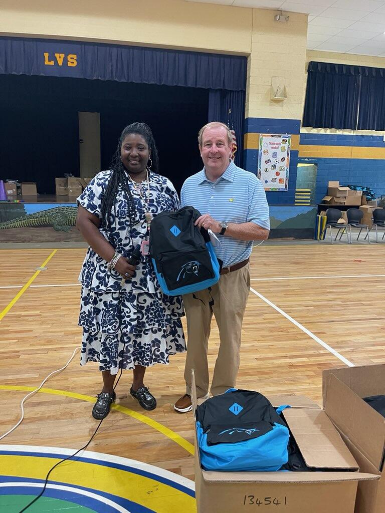 Two people stand in a gym holding backpacks, with more backpacks and boxes visible on the floor around them.