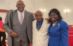 Three adults dressed formally stand together indoors, smiling at the camera.