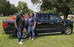 Two people stand smiling in front of a black pickup truck parked on grass, with trees and playground equipment visible in the background.