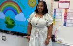 A woman stands in a classroom beside a smartboard displaying a rainbow and sun, with colorful charts on the wall.