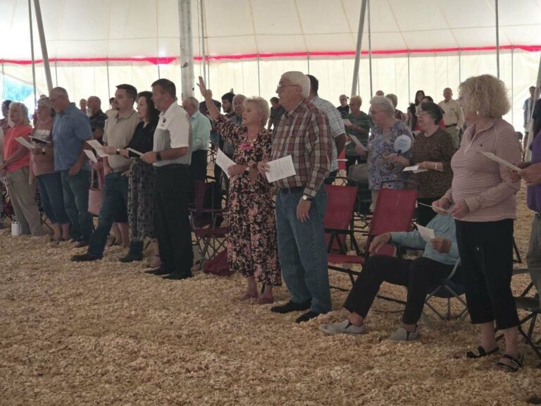 People standing and singing in a tent, holding papers, with chairs and wood chips on the ground.