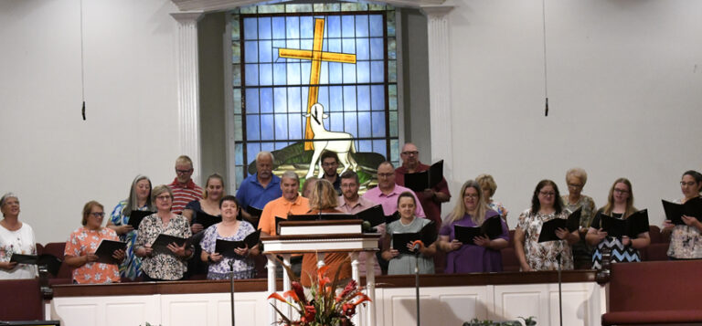 A church choir sings in front of a stained glass window depicting a cross and a lamb, with a podium and flowers in the foreground.