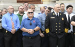 A group of men, including firefighters and officials, stand together outside a building, some in uniforms, posing for a photo.