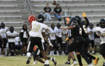 High school football players in black and white uniforms compete on the field, as the quarterback prepares to throw the ball. Spectators watch from the bleachers.