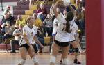Girls’ volleyball players in white uniforms compete in a gymnasium. One player prepares to set the ball, while spectators watch from the bleachers in the background.