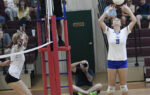 A volleyball player in blue prepares to set the ball as a teammate in white jumps. A referee and spectators watch in the background.