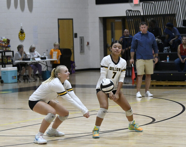 Two female volleyball players in white uniforms prepare to receive a serve in a gymnasium, with a coach and spectators visible in the background.