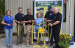 Five people stand together, smiling and holding large yellow ribbons and scissors at a ribbon-cutting ceremony in front of a sign for Midpoint I-95 RV Park.