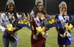 Three women wearing crowns and sashes hold bouquets of flowers while standing on a sports field at night during a pageant or ceremony.