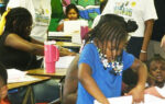 Children and adults engaged in activities at a classroom table, with colorful cartoon murals on the wall in the background.