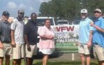 Four people stand together outdoors in front of a VFW Golf Tournament banner, smiling at the camera with trees and vehicles in the background.