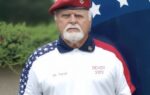 Older man with white beard wearing a red beret and patriotic shirt stands in front of a large American flag outdoors.