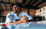 A man in a uniform and cap smiles while leaning on a vehicle inside a garage, with emergency vehicles visible in the background.