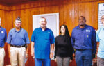 Four people standing indoors in front of a wooden-paneled wall, dressed in casual clothing, posing for a group photo.