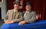 Two men wearing suits and hats smile while sitting at a blue table, with small American flags and a red curtain in the background.