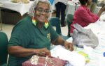 Smiling elderly woman with gray hair and colorful earrings sits at a table, working with fabric, in a community or craft setting.