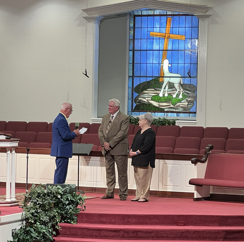 Three people stand at the front of a church with red pews and a stained glass window showing a cross and lamb.