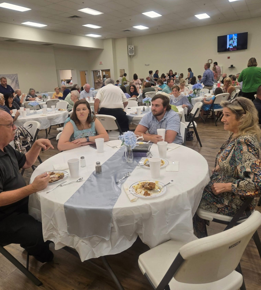 People sitting around decorated tables eating and talking at a large indoor event or gathering, with many attendees visible in the background.