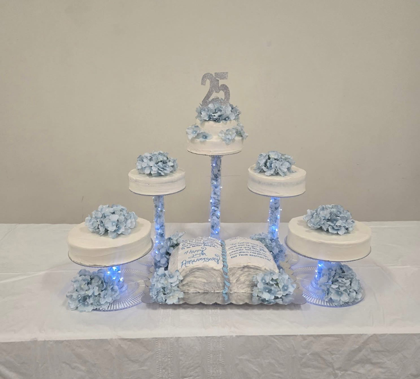 A tiered white and blue cake display with five cakes decorated with blue flowers and a "25" topper, arranged on a white table.