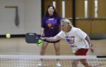 Two women playing pickleball indoors, one in a white shirt hitting the ball near the net, the other in a purple shirt standing behind her.