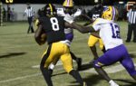 A football player in a black uniform runs with the ball while players in purple and yellow attempt to tackle him during a night game.