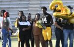A group celebrates on a football field, featuring a player in uniform, women holding flowers, and large gold balloon numbers. Bleachers are visible in the background.