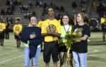 Four people are standing on a field at night, smiling, with one holding flowers and another holding a trophy, bleachers in the background.