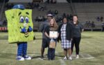 A group of people, including a mascot in a sponge costume, stand on a football field. One person holds a certificate, others are smiling.