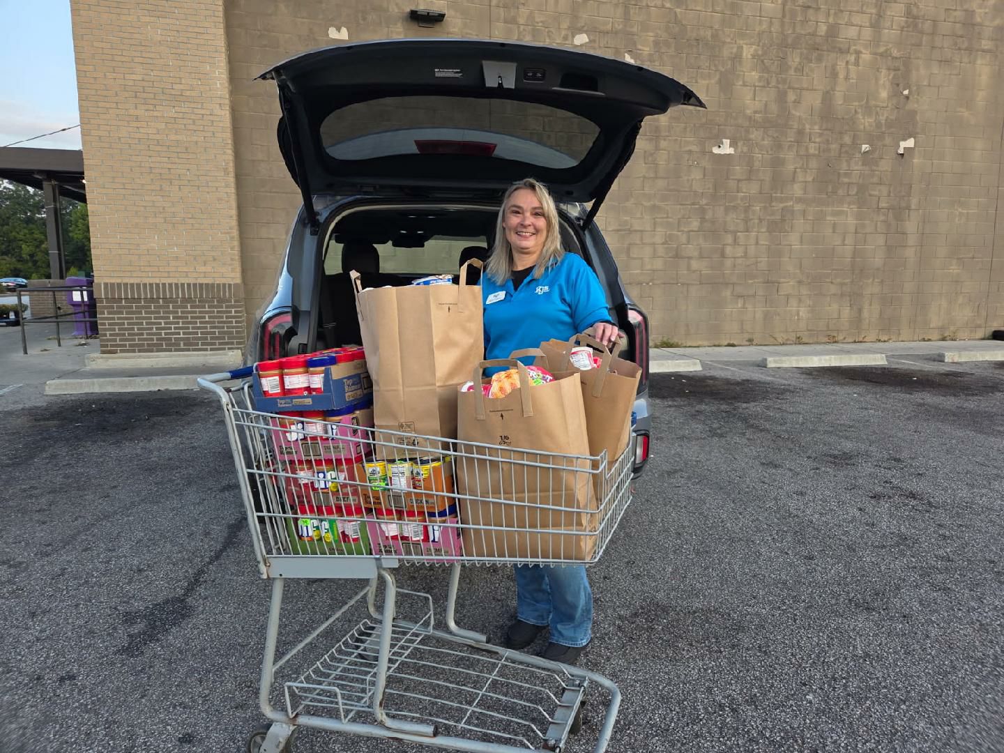 A woman stands by a car loading groceries from a shopping cart into the trunk in a parking lot.