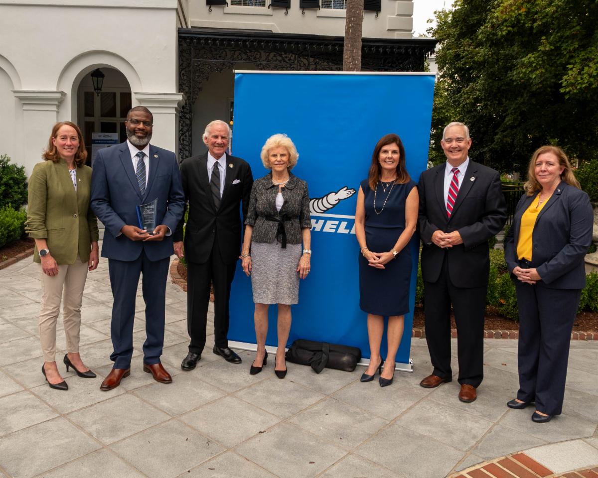 Seven people in business attire stand in a row outdoors, posing for a group photo in front of a blue sign and a white building.